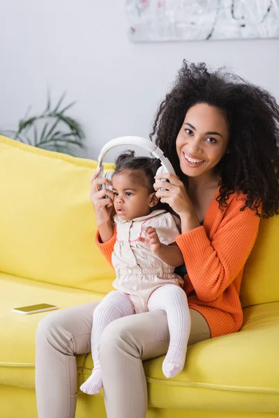 Heureuse mère afro-américaine portant des écouteurs sans fil sur la fille tout-petit — Photo de stock