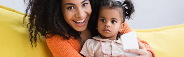 Madre afroamericana feliz sosteniendo teléfono inteligente cerca de niño pequeño, pancarta - foto de stock