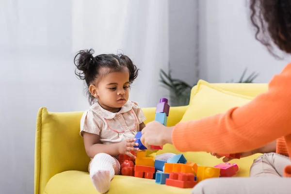 Africano americano bambino ragazza seduta su divano e giocare mattoni con la madre su sfocato primo piano — Foto stock