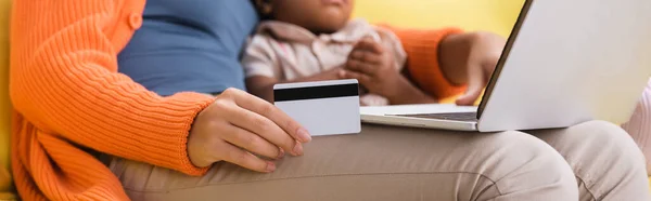 Vue partielle de la mère afro-américaine tenant une carte de crédit et utilisant un ordinateur portable près de la fille tout-petit, bannière — Photo de stock
