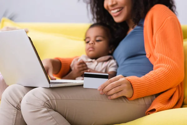 Feliz mãe afro-americana segurando cartão de crédito e usando laptop perto da filha da criança na sala de estar — Fotografia de Stock