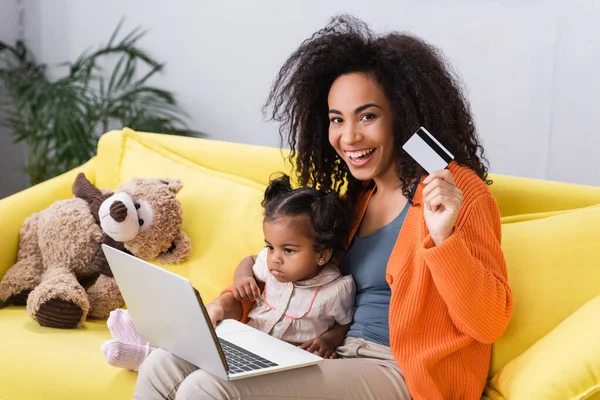 Sorrindo afro-americana mãe segurando cartão de crédito e usando laptop perto da filha da criança e brinquedo macio — Fotografia de Stock