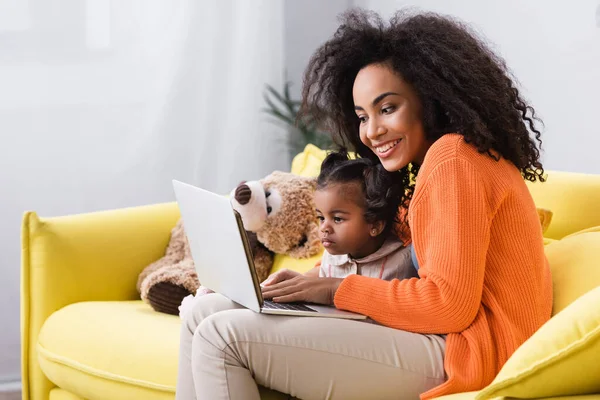 Усміхнена афроамериканська мати використовує ноутбук біля дочки малюка у вітальні — стокове фото