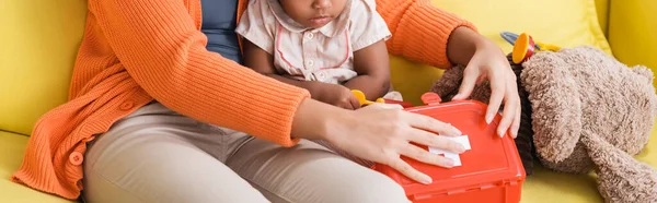 Visão recortada da mãe americana africana sentada com a filha da criança e segurando kit de primeiros socorros de brinquedo na sala de estar, banner — Fotografia de Stock