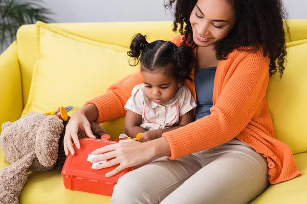 Happy african american mother sitting with toddler daughter and holding toy first aid kit in living room — Stock Photo