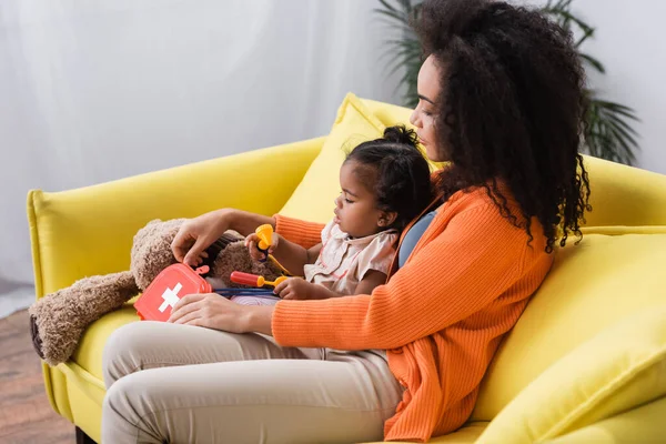 Madre afroamericana sentada con su hija pequeña y sosteniendo el kit de primeros auxilios de juguete en la sala de estar - foto de stock