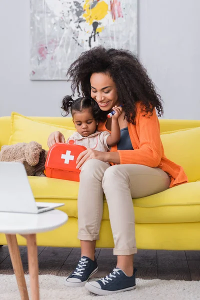 Felice madre afro-americana seduta con la figlia del bambino e con in mano il kit di primo soccorso giocattolo in soggiorno — Foto stock