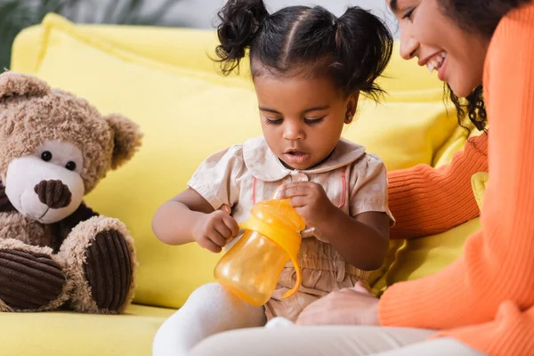 Happy african american mother looking at toddler daughter with baby bottle — Stock Photo