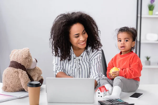 Happy african american mother looking at toddler daughter sitting on desk with apple near gadgets — Stock Photo