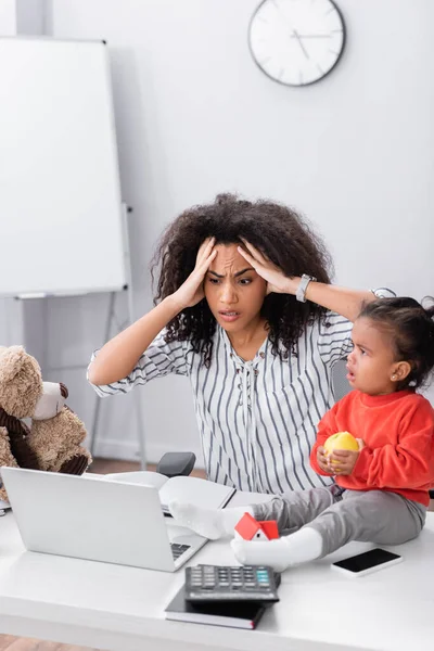 Stressé afro-américain freelance près de tout-petit fille assis sur le bureau avec pomme — Photo de stock