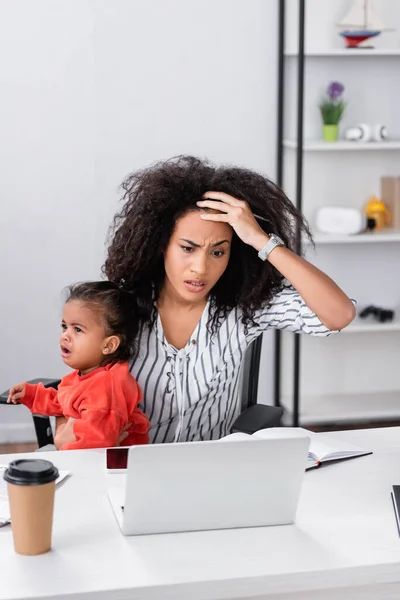 Estresado afroamericano freelancer sosteniendo en brazos llorando hija pequeño mientras trabaja desde casa - foto de stock