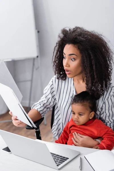 Überrascht afrikanisch-amerikanische Mutter hält Ordner in der Hand, während sie mit ihrer kleinen Tochter sitzt und von zu Hause aus arbeitet — Stockfoto
