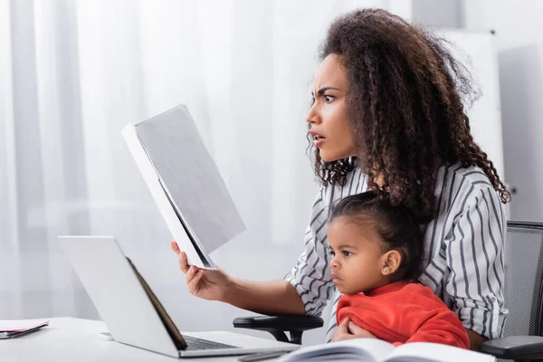 Gestresste afrikanisch-amerikanische Mutter hält Ordner in der Hand, während sie mit ihrer kleinen Tochter sitzt und von zu Hause aus arbeitet — Stockfoto