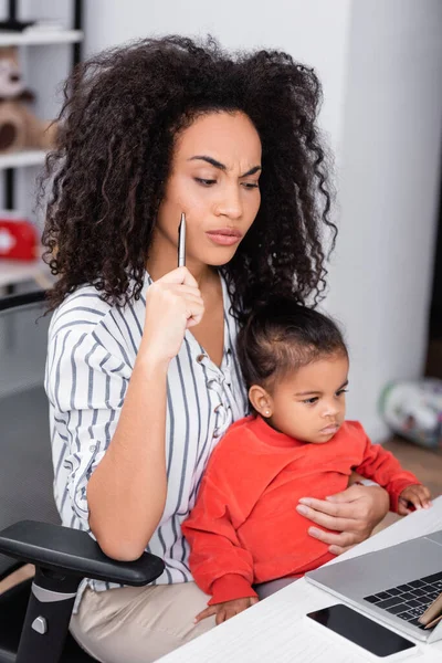 Nachdenkliche afrikanisch-amerikanische Mutter hält Stift in der Hand und sitzt mit ihrer kleinen Tochter, während sie von zu Hause aus arbeitet — Stockfoto
