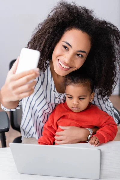 Feliz afro-americana mãe tomando selfie com a filha da criança perto do laptop em primeiro plano borrado — Fotografia de Stock
