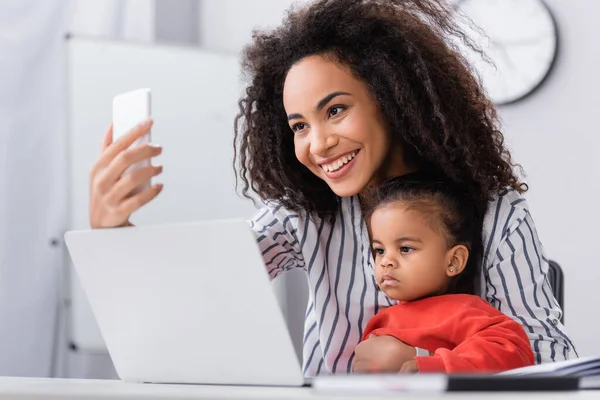 Щаслива афроамериканська мати бере селфі з дочкою біля ноутбука на розмитому передньому плані — стокове фото