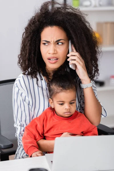 African american mother talking on smartphone while sitting with toddler daughter near laptop on blurred foreground — Stock Photo