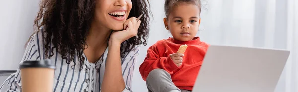 Tout-petit enfant afro-américain tenant un biscuit savoureux près de la mère heureuse travaillant de la maison, bannière — Photo de stock