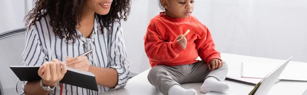 Toddler african american kid holding tasty cookie and sitting on desk near mother writing in notebook near laptop, banner — Stock Photo
