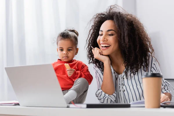 Niño afroamericano niño sosteniendo sabrosa galleta y sentado en el escritorio cerca de madre feliz mirando portátil - foto de stock