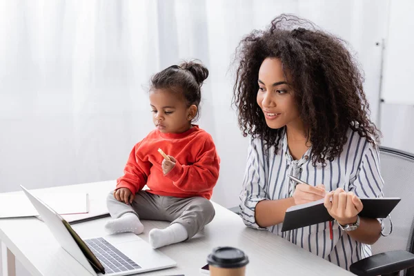 Kleinkind afrikanisch-amerikanisches Kind hält leckeren Keks in der Hand und sitzt auf dem Schreibtisch neben der Mutter, die von zu Hause aus arbeitet — Stockfoto