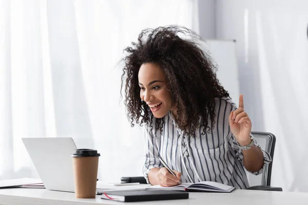 Aufgeregte Afroamerikanerin schreibt in Notizbuch und schaut auf Laptop, während sie mit dem Finger zeigt — Stockfoto