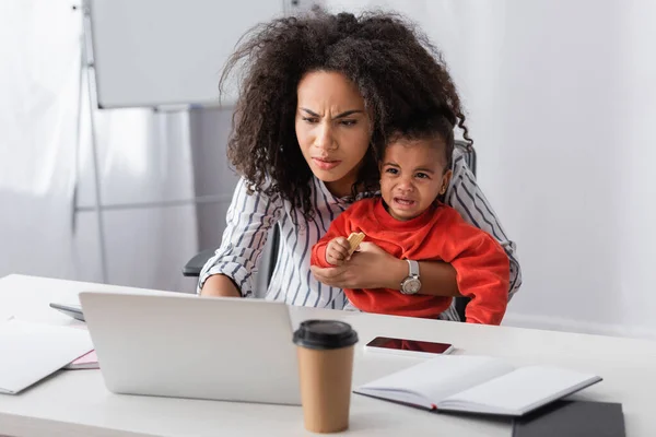 Gestresste afrikanisch-amerikanische Mutter hält weinendes Kleinkind in den Armen, während sie von zu Hause aus arbeitet — Stockfoto