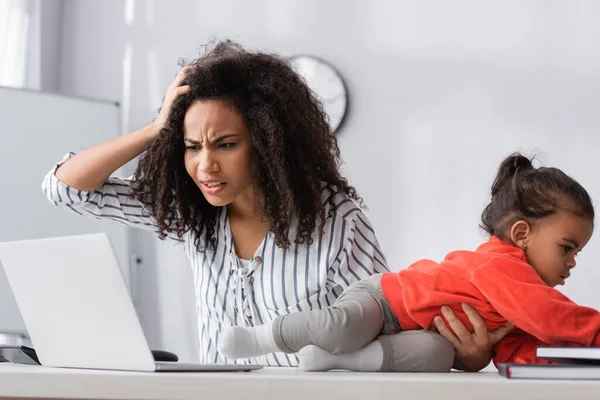 A souligné la mère afro-américaine ajustant les cheveux et regardant l'ordinateur portable près de tout-petit enfant rampant sur le bureau — Photo de stock