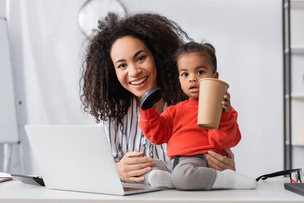 Малюк африканський американець сидить на столі і тримає паперову чашку біля веселої матері. — стокове фото