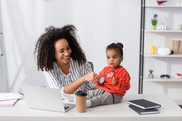 Glückliche afrikanisch-amerikanische Mutter schaut Tochter mit Brille an, während sie auf dem Schreibtisch sitzt — Stockfoto