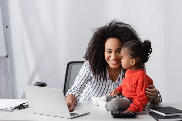 Улыбающаяся африканская американская мать смотрит на дочь малыша, сидящую рядом с ноутбуком на столе — стоковое фото