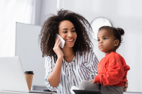 Усміхнена афроамериканська мати розмовляє по смартфону і дивиться на дочку малюка на розмитому передньому плані — стокове фото