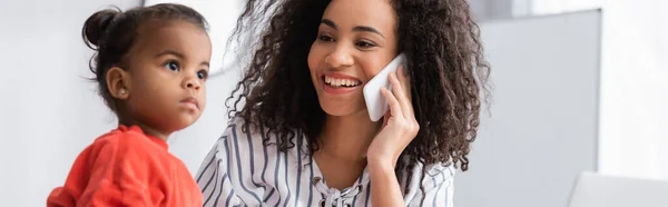 Sonriente madre afroamericana mirando a la hija del niño pequeño mientras habla en el teléfono inteligente, pancarta - foto de stock