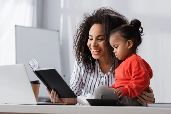 Lächelnde afrikanisch-amerikanische Mutter schaut auf Notizbuch in der Nähe von Kleinkind Tochter auf dem Schreibtisch — Stockfoto