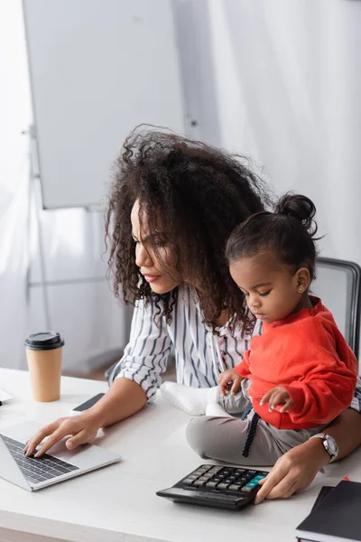 Focused african american mother using laptop toddler daughter on desk — Stock Photo