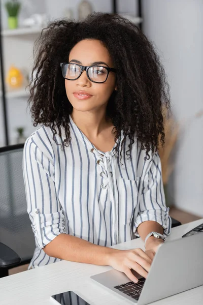 Afro-américaine pigiste dans des lunettes détourner les yeux et en utilisant un ordinateur portable à la maison — Photo de stock