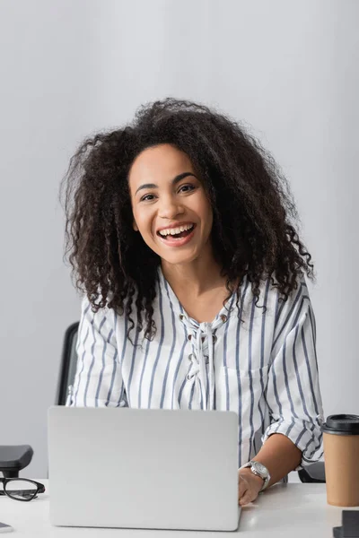 Feliz afroamericano freelancer sonriendo mientras mira la cámara cerca del ordenador portátil en el escritorio - foto de stock