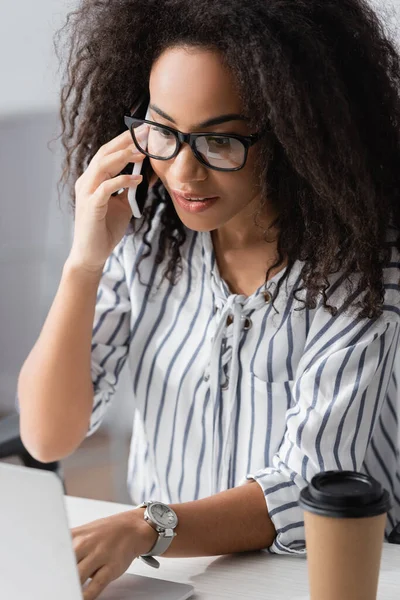 African american freelancer in glasses talking on smartphone near laptop at home — Stock Photo