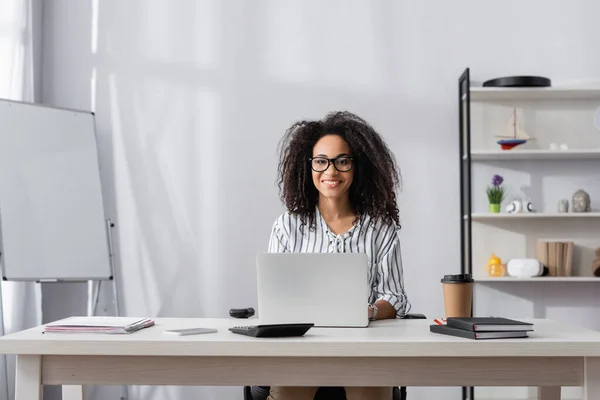 Joyeux freelance afro-américain dans des lunettes à l'aide d'un ordinateur portable près du smartphone, ordinateurs portables et tasse en papier sur le bureau — Photo de stock