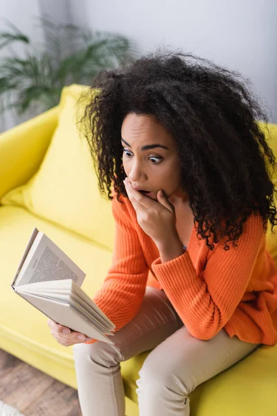 Shocked african american woman reading book while sitting on couch — Stock Photo