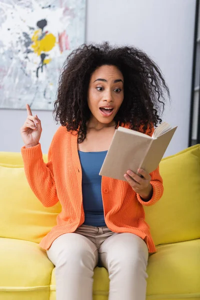 Mujer afroamericana asombrada teniendo idea al leer libro y sentada en el sofá - foto de stock