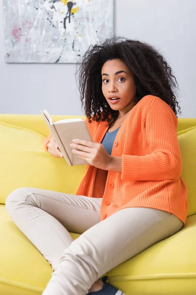 Surprised african american woman holding book while sitting on couch — Stock Photo