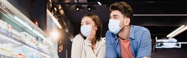 Молодая пара в медицинских масках в супермаркете, баннер — стоковое фото