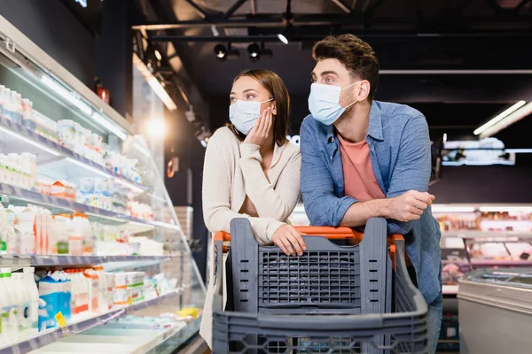 Pareja en máscaras médicas mirando comestibles cerca de carrito de compras en el supermercado - foto de stock
