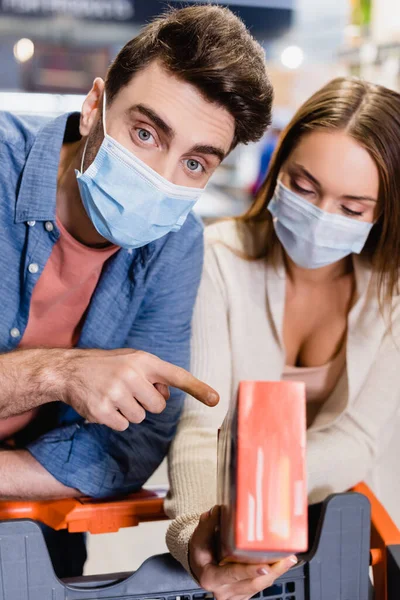 Man in medical mask pointing at box near girlfriend and shopping trolley in supermarket — Stock Photo