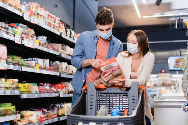 Couple in medical masks looking at box near shopping trolley on blurred foreground in store — Stock Photo