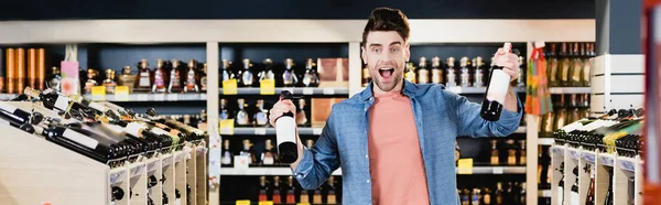 Взволнованный человек держит бутылки вина в супермаркете, баннер — стоковое фото