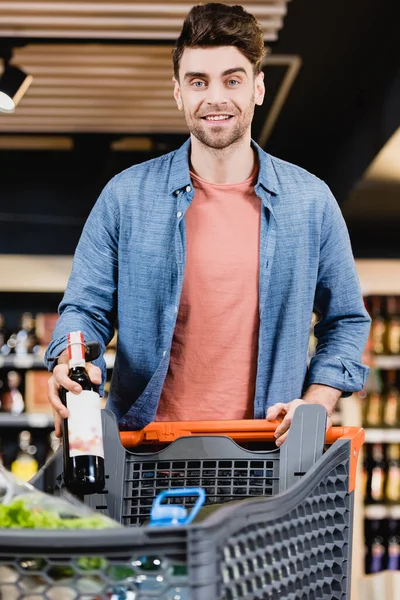 Homme souriant avec bouteille de vin regardant la caméra près du panier en magasin — Photo de stock