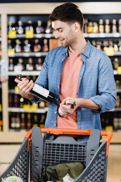 Homme joyeux tenant des bouteilles de vin près du chariot en magasin — Photo de stock