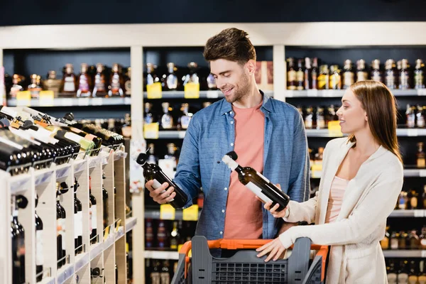 Casal jovem sorrindo enquanto segurava garrafas de vinho perto do carrinho de compras no supermercado — Fotografia de Stock